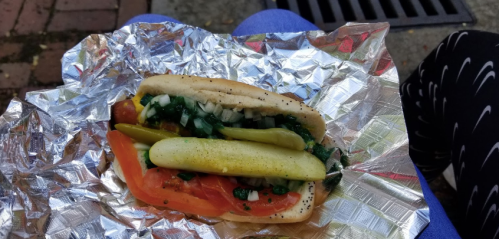 A hot dog in foil, topped with tomatoes, pickles, onions, and green relish, resting on a person's lap.