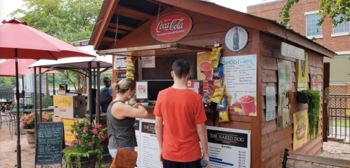 A wooden food stand with a Coca-Cola sign, serving smoothies and snacks, with customers ordering at the counter.