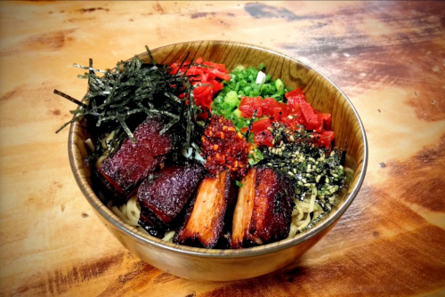 A bowl of ramen topped with sliced pork, seaweed, green onions, and diced vegetables on a wooden surface.