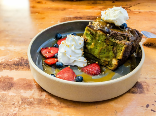 A slice of green cake topped with whipped cream, surrounded by strawberries, blueberries, and caramel sauce on a plate.