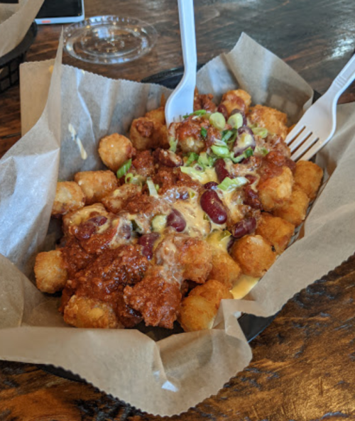 A basket of tater tots topped with chili, cheese, and green onions, served with two plastic forks.