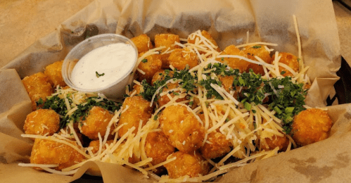 A basket of golden tater tots topped with shredded cheese and herbs, served with a side of ranch dressing.