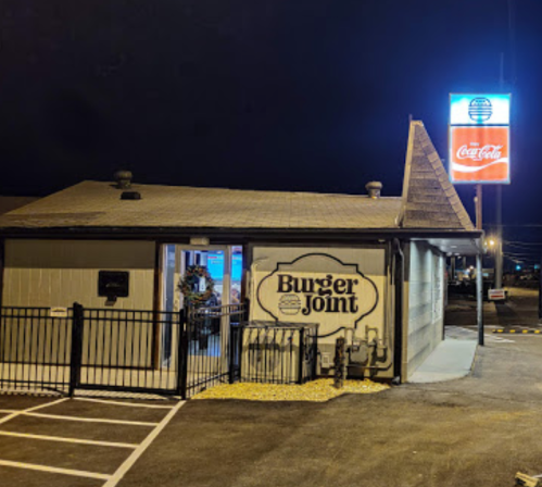 A nighttime view of a Burger Joint with a lit sign and a fenced outdoor area.