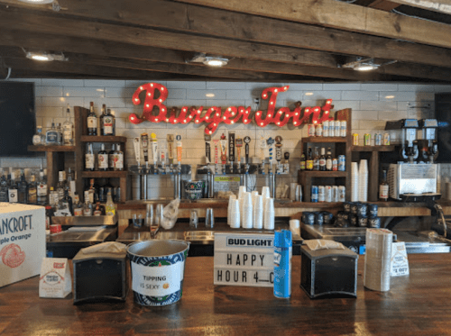 A bar counter with various liquor bottles, cups, and a neon "Burger Joint" sign in a rustic setting.