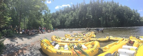 A group of people by a river with yellow rafts lined up on the shore, surrounded by trees and a clear blue sky.