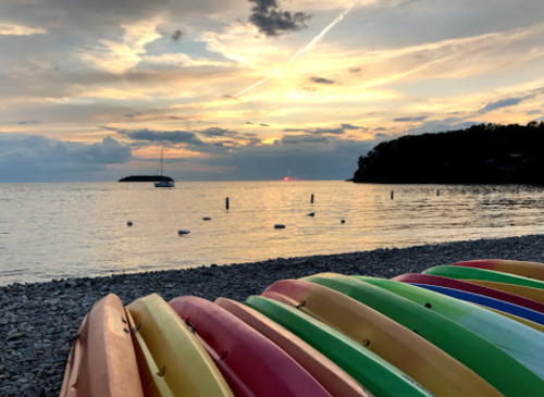 Colorful kayaks stacked on a rocky beach at sunset, with calm water and a distant sailboat under a cloudy sky.