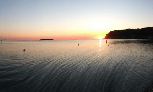 A serene sunset over calm waters, with a distant island and a silhouette of a shoreline.