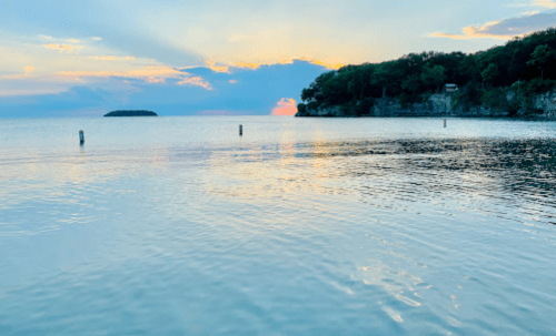 Serene lake at sunset, with calm waters reflecting the colorful sky and distant trees along the shoreline.