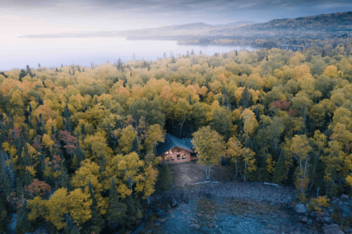 Aerial view of a cozy cabin surrounded by vibrant autumn trees near a serene lake.