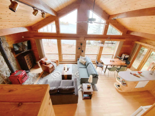 Cozy wooden cabin interior with large windows, a stone fireplace, and a comfortable living area overlooking nature.