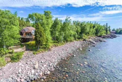 A scenic view of a rocky shoreline with a house surrounded by trees and clear blue water under a bright sky.