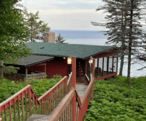 A red cabin surrounded by greenery, with a wooden path leading to it and a view of the lake in the background.