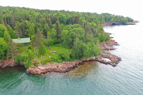 Aerial view of a lush green coastline with rocky shores and a cabin surrounded by trees near calm waters.