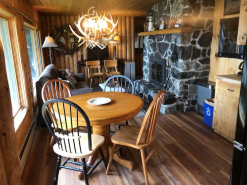 Cozy cabin interior featuring a wooden dining table, chairs, stone fireplace, and large windows with natural light.