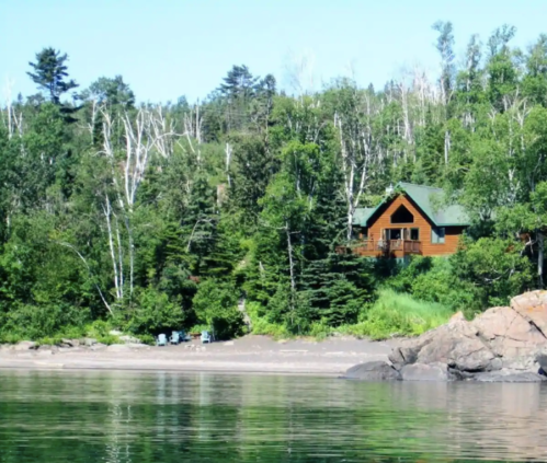A wooden cabin nestled among lush green trees by a calm lake, with a rocky shore in the foreground.