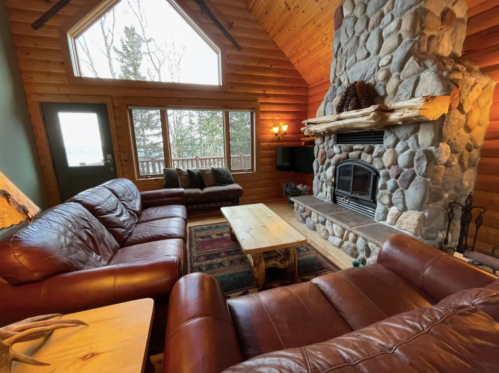 Cozy cabin living room with leather sofas, a stone fireplace, and large windows overlooking a forested view.