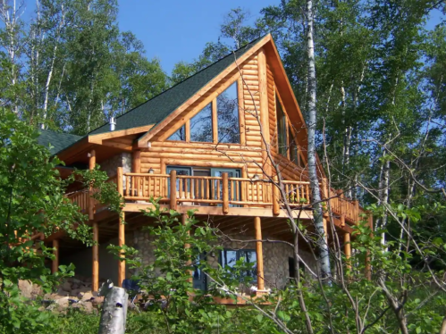 A large log cabin with a green roof, surrounded by trees and a clear blue sky.