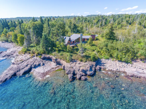 Aerial view of a cabin by a rocky shoreline, surrounded by lush greenery and clear blue water.