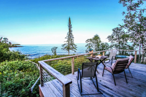 A scenic deck overlooking the ocean, surrounded by trees and greenery, with two chairs and a table set for relaxation.