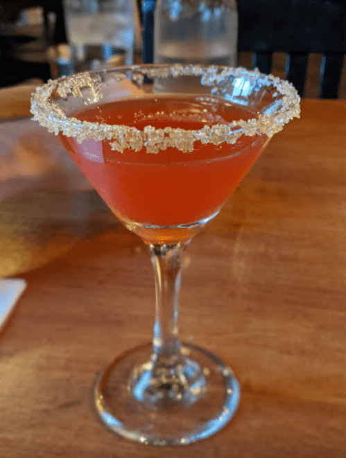 A vibrant pink cocktail in a glass with a sugar rim, sitting on a wooden table.