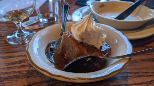 A slice of dessert topped with whipped cream, served on a plate with spoons and remnants of a drink in the background.