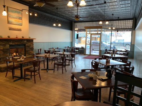 Cozy restaurant interior with wooden tables, chairs, a fireplace, and large windows letting in natural light.