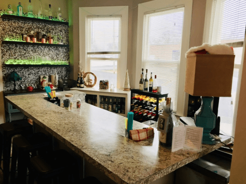 A stylish home bar with a granite countertop, shelves of bottles, and festive decorations in a bright room.