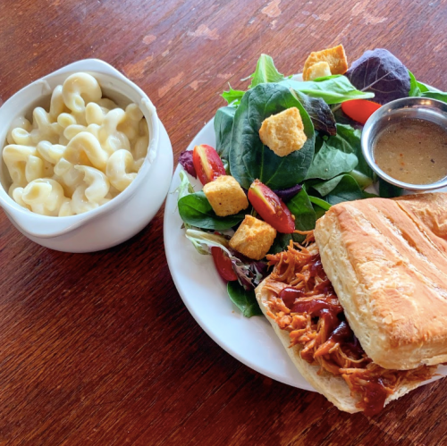 A plate with a BBQ chicken sandwich, side salad with croutons, and a bowl of macaroni and cheese.