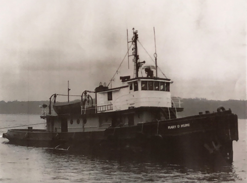 Black and white image of the tugboat "Mary D. Hume" on calm waters, with a forested shoreline in the background.
