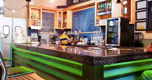A vibrant café bar with a wooden counter, shelves of drinks, and a person preparing coffee in the background.