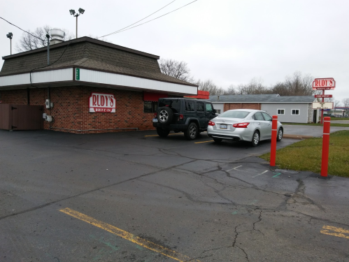 A brick building with a sign reading "Rudy's," featuring a black SUV and a white car parked outside.