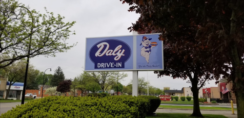 Sign for Daly Drive-In featuring a cartoon character holding a burger, surrounded by trees and a cloudy sky.