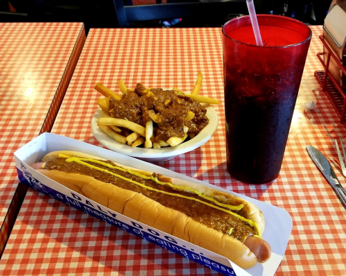 A hot dog with mustard, a plate of chili cheese fries, and a drink on a checkered tablecloth.