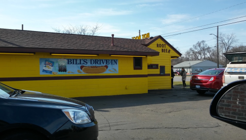 A bright yellow drive-in restaurant with a sign for root beer and a banner reading "Bill's Drive-In."
