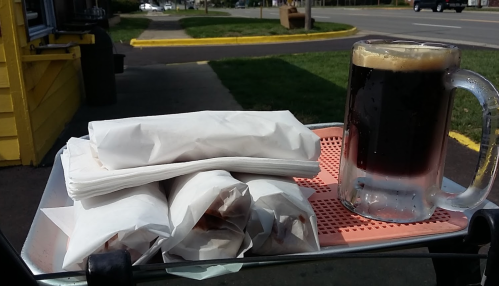 A tray with wrapped food items and a frosty mug of dark beverage, set outdoors near a road.