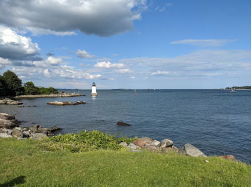 A scenic view of a lighthouse by the water, with rocky shorelines and a blue sky dotted with clouds.