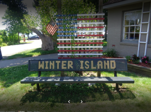 A bench with "WINTER ISLAND" engraved, set against a backdrop of an American flag and greenery.