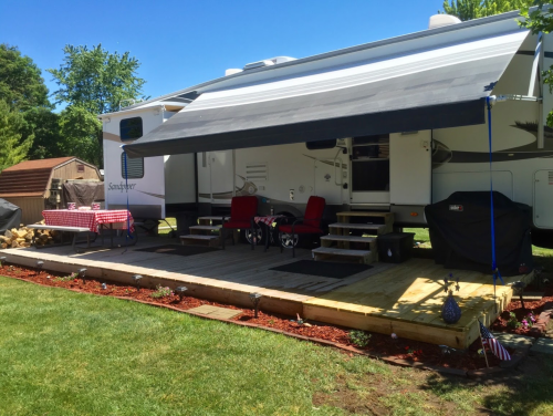 A parked RV with an awning, surrounded by a wooden deck, picnic table, and lawn in a sunny outdoor setting.