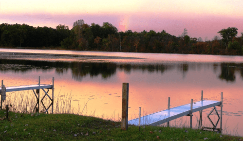 A serene lake at sunset, with a wooden dock extending into calm waters reflecting colorful skies and surrounding trees.