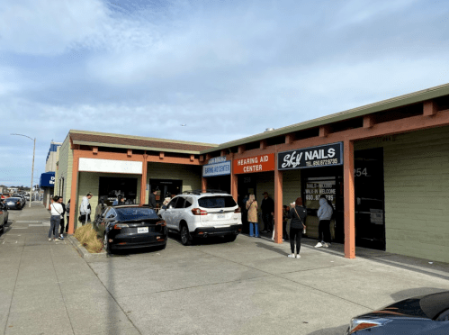 A busy street scene showing a hearing aid center and a nail salon, with people waiting outside and parked cars.