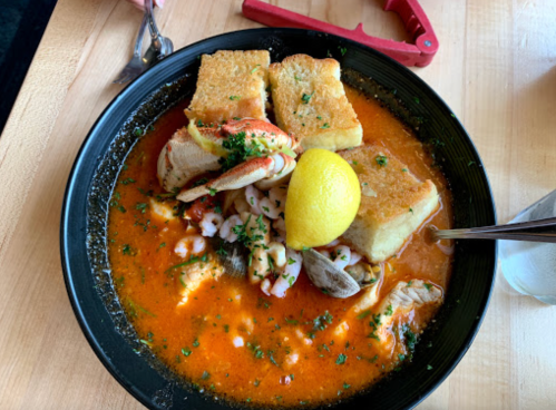 A bowl of seafood stew with bread, lemon, and herbs, featuring shrimp, clams, and other seafood in a rich broth.