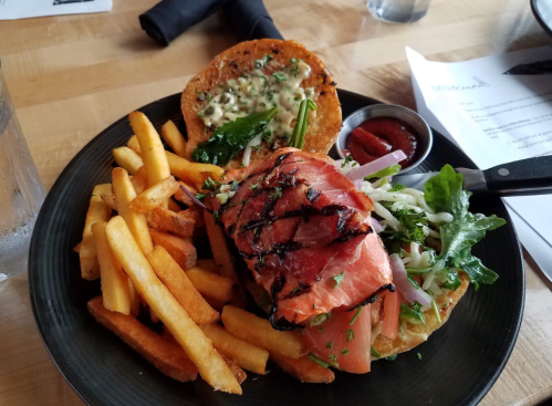 A grilled salmon sandwich with lettuce and tomato, served with fries and a side of ketchup on a black plate.