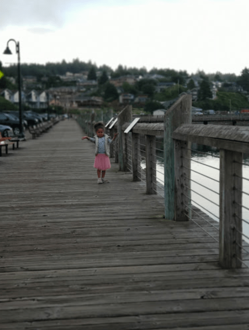 A child in a pink skirt stands on a wooden pier, with water and trees in the background.