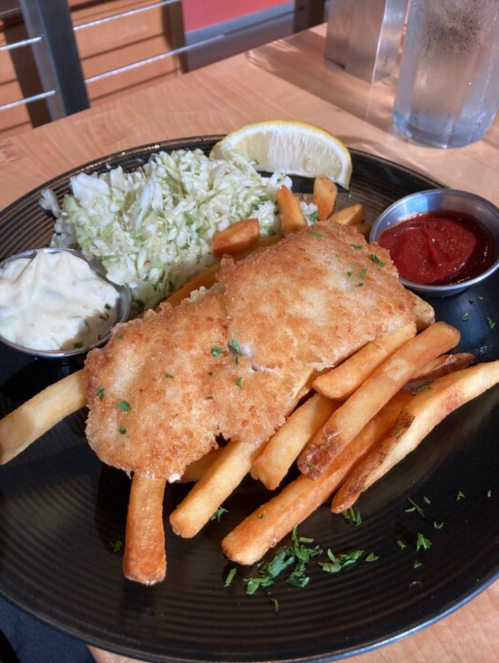 A plate of crispy fried fish fillets served with French fries, coleslaw, a lemon wedge, and dipping sauces.