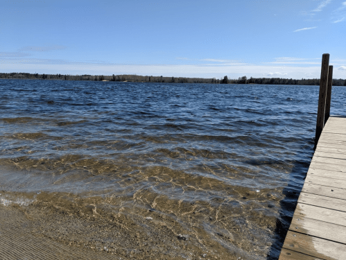 A calm lake with clear water and a wooden dock under a blue sky, surrounded by trees in the distance.