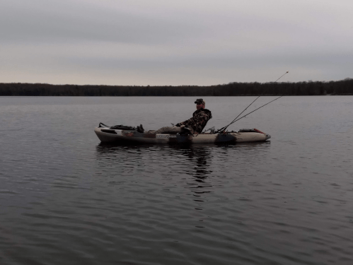 A person fishing from a kayak on a calm lake, surrounded by trees under a cloudy sky.