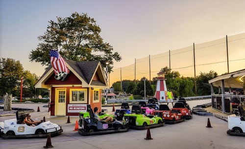 Go-kart track with colorful karts parked near a ticket booth and a lighthouse, under a sunset sky.