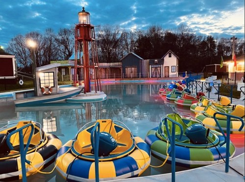 Colorful bumper boats lined up in a pool, with a lighthouse and rustic buildings in the background under a cloudy sky.