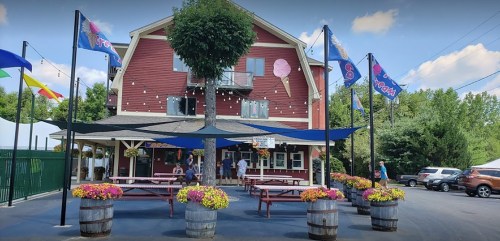 A red building with flags and flowers, featuring outdoor seating and people enjoying a sunny day.