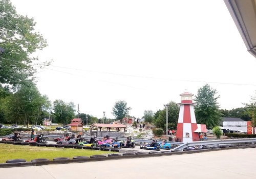 A go-kart track with colorful karts and a lighthouse in the background, surrounded by trees and a parking area.